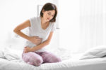 Young woman suffering from abdominal pain while sitting on bed at home.